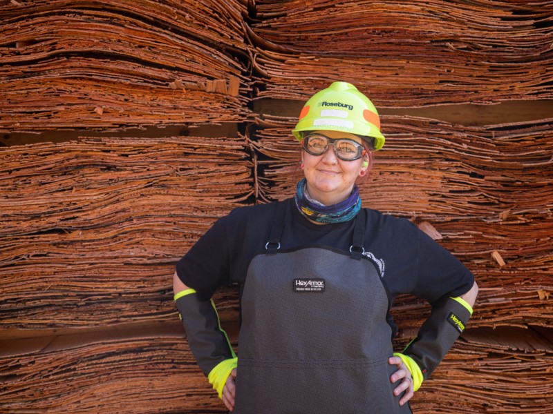 A female Roseburg team member stands in front of two tall stacks of veneer