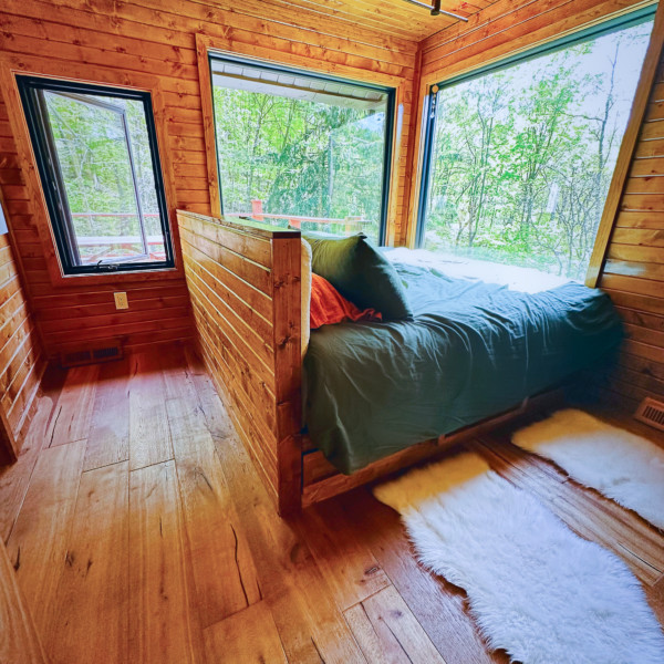 Interior view of a bedroom covered in floor-to-ceiling wood. A bed sits in front of two picture windows.